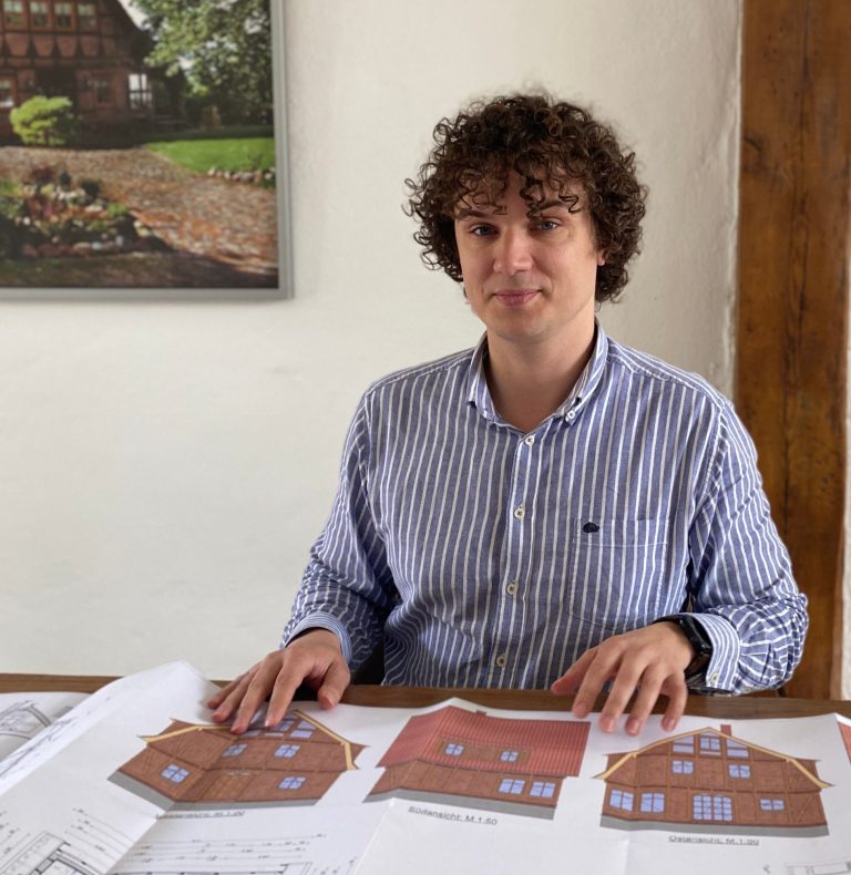 Björn Christianus Mann mit lockigem Haar sitzt an einem Tisch mit Bauplänen von Häusern.