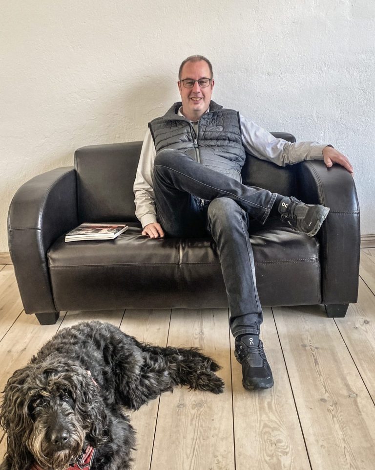 Hendrik Paulsen Mann sitzt auf einem Sofa, ein Hund liegt entspannt neben ihm.